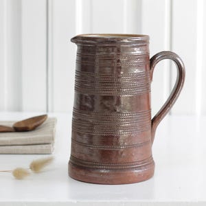 Puede incluir: Jarra de cerámica marrón con asa y boquilla. La jarra tiene bandas horizontales texturizadas alrededor del cuerpo. Una cuchara de madera y un paño doblado están en el fondo.