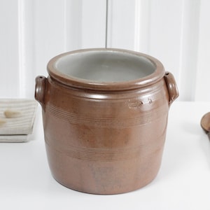 May include: A brown ceramic pot with two handles and horizontal lines. A wooden spoon rests nearby. The pot has a light interior and sits on a white surface. A folded linen cloth is also present.