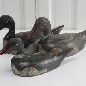 May include: Three carved wooden duck decoys featuring weathered dark paint with visible red and white accents on the bills and bodies. Each duck has a distinct head shape and posture, showing signs of age and wear on the painted surfaces.