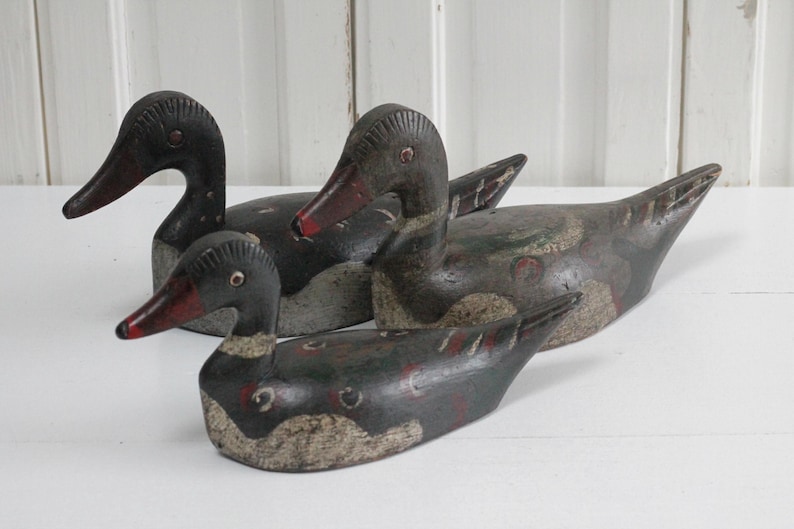 May include: Three carved wooden duck decoys featuring weathered dark paint with visible red and white accents on the bills and bodies. Each duck has a distinct head shape and posture, showing signs of age and wear on the painted surfaces.