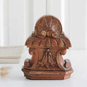 May include: A detailed, antique-style wooden decorative element. The piece is a warm reddish-brown color, featuring ornate carvings of leaves and scrolls. The top has a shell-like design. The item is likely a decorative architectural piece or a small box.