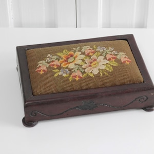 May include: A rectangular dark brown wooden box featuring a needlepoint insert on the lid. The needlepoint displays a floral arrangement with pink, yellow, and green threads on a brown background. The box has carved scrollwork and four feet.