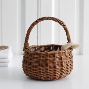 May include: A round, woven brown wicker basket with a handle. The basket has a small, fluffy, light-colored accent on the side. A stack of white folded towels and a wooden spatula are in the background.