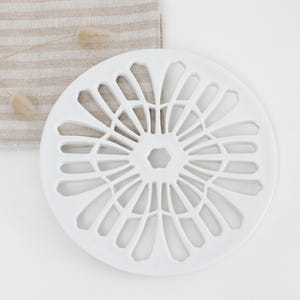 May include: A white, circular trivet with a floral design. The trivet has a central hexagonal shape surrounded by petal-like cutouts. It sits on a white surface with a beige and white striped cloth and dried flowers in the background.