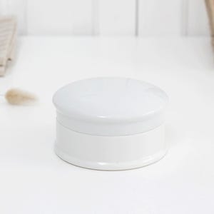 May include: A white ceramic jar with a rounded lid and a cylindrical base. The jar is sitting on a white surface. The background includes a white wall, a woven basket, and a folded beige towel.