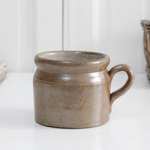 May include: A short, wide-bodied ceramic mug with a glossy, light brown speckled glaze. The mug features a thick, rounded rim, a straight cylindrical base, and a single curved handle attached to the side, showcasing a smooth, reflective finish.