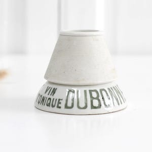 May include: A white ceramic object with a tapered, ribbed upper section and a wider, smooth base. The base features the dark green printed text VIN TONIQUE DUBONNET. The object has a hollow centre and a flared, circular opening at the top.