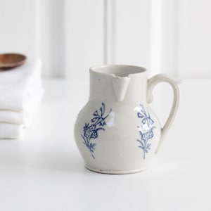 May include: A small, off-white ceramic jug with a handle and spout. The jug is decorated with blue floral designs. A wooden spoon and a stack of white towels are in the background.