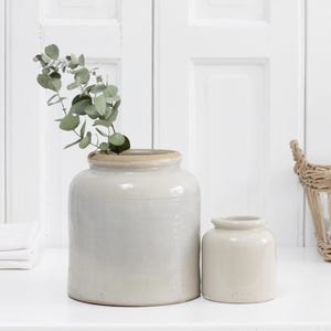 May include: Two cylindrical ceramic crocks with a smooth, off-white glazed finish. The larger crock on the left contains a sprig of green eucalyptus leaves. Both vessels feature a simple, wide-mouthed design with a subtle rim at the top opening.