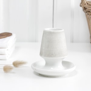 May include: A white ceramic candle holder featuring a tapered, ribbed upper section and a smooth, circular base. The design incorporates a flared neck connecting the textured top to the wide, flat bottom, creating a singular, cohesive structure.