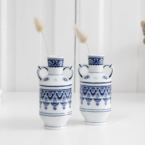 Może przedstawiać: Dwie białe ceramiczne wazony z niebieskimi wzorami dekoracyjnymi, każdy z dwoma bocznymi uchwytami i wąską szyjką. W każdym wazonie umieszczono pojedynczą łodygę suszonej beżowej trawy, wazon ma cylindryczny korpus i rozszerzaną podstawę.