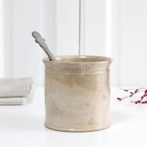 Può includere: Un portautensili in ceramica beige con un cucchiaio d'argento all'interno. Il portautensili ha una forma cilindrica con un bordo e una base leggermente svasati. Sono visibili anche una pila di asciugamani beige piegati e un rametto di bacche rosse.