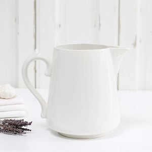 May include: A plain white ceramic jug with a curved handle and a pouring spout. The jug has a smooth, glossy finish and a wide base. It is positioned next to a stack of folded white linens and a small bundle of dried purple lavender stems.