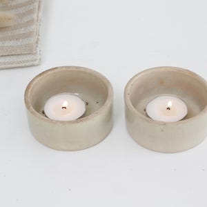 Könnte beinhalten: Zwei kleine, beige Keramik-Kerzenhalter, die jeweils ein brennendes weißes Teelicht enthalten. Die Halter sind rund und haben eine leicht raue Textur. Im Hintergrund befindet sich ein gefalteter, gestreifter Stoff.