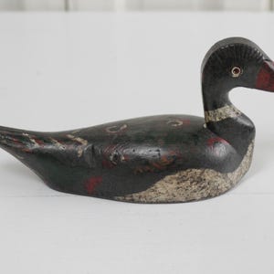May include: A hand-carved wooden duck decoy featuring a black body with distressed paint, a white textured chest, and a red-tipped beak. The figure has a carved eye detail and a slender neck, showcasing a weathered finish with visible wood grain.