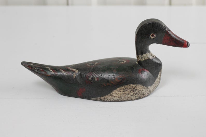 May include: A hand-carved wooden duck decoy featuring a black body with distressed paint, a white textured chest, and a red-tipped beak. The figure has a carved eye detail and a slender neck, showcasing a weathered finish with visible wood grain.