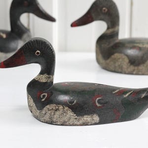 May include: Three carved wooden duck decoys featuring dark black, green, and red painted details. Each duck has a distinct shape with a pointed tail, a curved neck, and a beak with red accents. The paint shows signs of wear and texture.