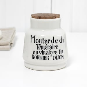 以下が含まれることがあります： コルクの蓋が付いた白いセラミックジャー。ジャーには「Moutarde du Téméraire au vinaigre fin BORNIER DIJON」と書かれた黒い文字があります。ジャーは白い表面にあります。