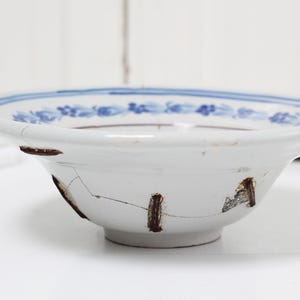 May include: A white ceramic bowl with blue and brown floral designs around the rim. The bowl has several cracks and chips, giving it a vintage look.