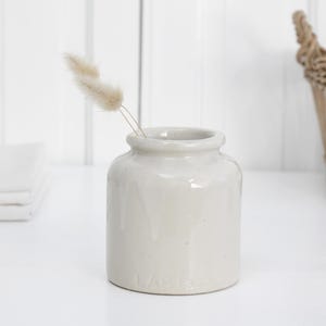 May include: A small, off-white ceramic vase with a rounded body and a narrow neck, holding two dried, fluffy stems. The vase sits on a white surface, with a wicker basket and folded white linens in the background. The vase is approximately 10 cm tall.