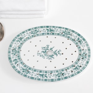 May include: An oval, white ceramic serving dish with a green plaid and floral border. The dish has a central hole and small holes throughout. A silver spoon and folded white linens are also present.