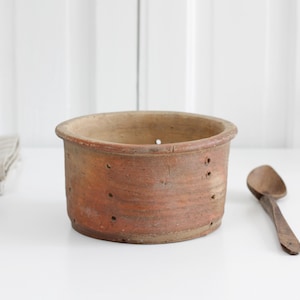 Könnte beinhalten: Ein rustikaler, zylindrischer Käseform aus Terrakotta mit kleinen Löchern, neben einem Holzlöffel. Die Form hat einen breiten Rand und ein verwittertes Aussehen. Ein Stapel gefalteter Leinentücher befindet sich links.