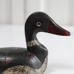 May include: A hand-carved wooden duck decoy featuring a black body with distressed white painted details on the chest and neck. The head has a dark red beak, a textured crest, and a painted eye with a red iris. The surface shows signs of wear.