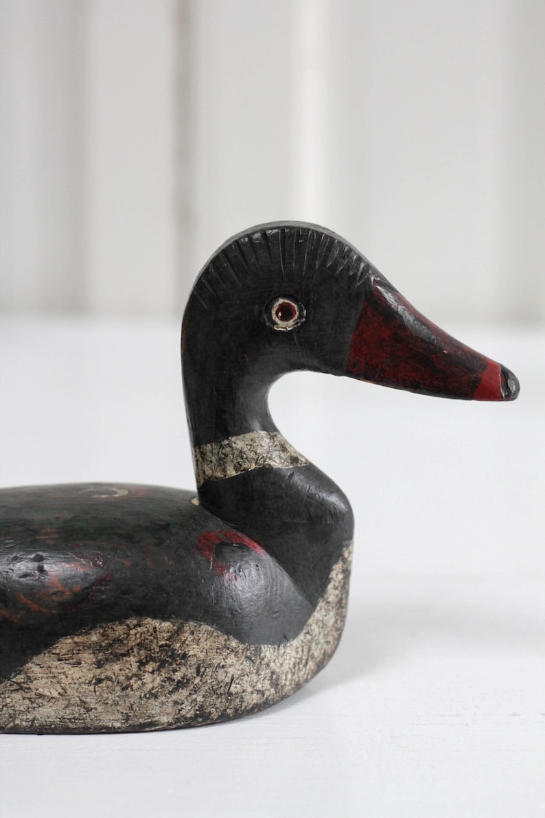 May include: A hand-carved wooden duck decoy featuring a black body with distressed white painted details on the chest and neck. The head has a dark red beak, a textured crest, and a painted eye with a red iris. The surface shows signs of wear.