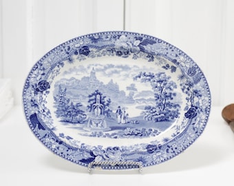 Op de afbeelding: Ovale blauwe en witte keramische schaal met een landschapsontwerp. De rand heeft bloemen- en scrollpatronen. De schaal staat op een metalen standaard, met een houten lepel rechts. Ongeveer 30 cm lang.