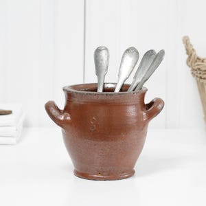 May include: A brown glazed earthenware pot with two side handles, containing four silver-toned metal spoons. The pot has a rounded body and a wide opening, displaying a smooth, glossy finish on the exterior surface and a simple, functional design.