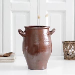 May include: A brown ceramic vase with two handles. The vase has a glossy finish and sits on a white surface. A woven basket and a wooden spoon are also visible in the image. The vase is a decorative item.