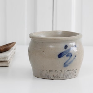 May include: A light grey stoneware crock with a rounded body and a wide opening. The front features a hand-painted blue mark above the word MARGARINE embossed in capital letters. The surface has a smooth, matte finish with subtle speckling.