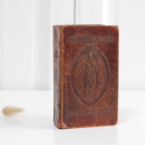 Puede incluir: Un libro antiguo de cuero marrón con imágenes religiosas en relieve. El libro presenta una ilustración detallada dentro de un marco ovalado en la portada. El lomo tiene texto y el libro está sobre un fondo blanco.