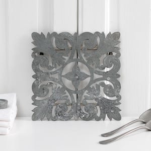 Puede incluir: Un adorno de pared decorativo de metal gris con un diseño ornamental y simétrico. Dos cucharas a juego y un pequeño recipiente redondo se encuentran cerca de una pila de toallas blancas. El adorno de pared mide unos 30 cm cuadrados.