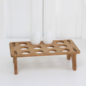 May include: A rectangular light brown wooden egg holder stand featuring twelve circular cutouts arranged in three rows of four. Two smooth white oval-shaped eggs are positioned on the top surface of the wooden rack, which is supported by four short legs.