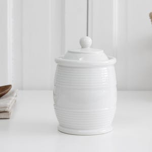 Może przedstawiać: Biały ceramiczny pojemnik z pasującą pokrywką z małym kulistym uchwytem. Cylindryczny korpus posiada szereg poziomych żłobień otaczających górną i dolną część, co tworzy subtelny wzór o wyczuwalnej fakturze na gładkiej powierzchni naczynia.