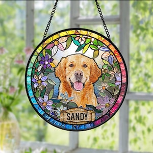 May include: A round stained glass window featuring a Golden Retriever dog with a happy expression, surrounded by colorful flowers. The dog's name, "Sandy," is displayed on a banner at the bottom. The window is framed in black and hangs from a chain.