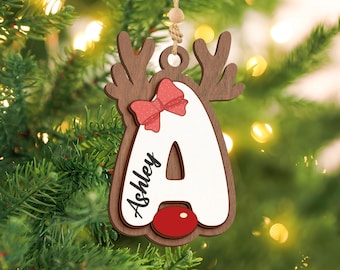 May include: A wooden Christmas ornament shaped like a reindeer with the letter "A" in white. The ornament has brown antlers, a red bow, and a red nose. The name "Ashley" is written on the ornament.