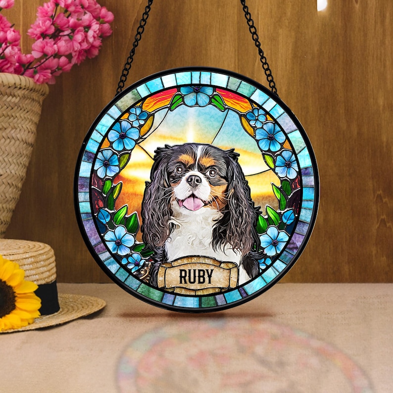 May include: Round stained glass window featuring a Cavalier King Charles Spaniel with the name "RUBY" on a banner. The window has a blue floral border and a colorful sunset background, hanging from a black chain.