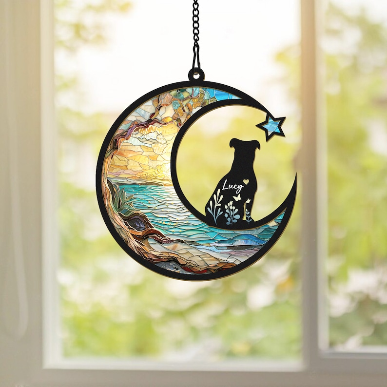 Pode incluir: Um apanhador de sol de vidro colorido em forma de lua crescente com uma silhueta de um c&atilde;o sentado dentro. A lua tem um design de mosaico com tons de azul, verde, amarelo e laranja. O c&atilde;o &eacute; preto e o texto "Lucy" est&aacute; escrito em branco dentro da lua.