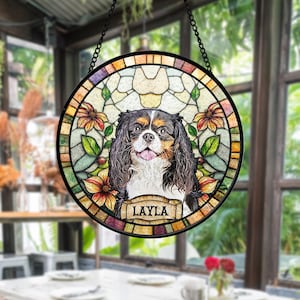 May include: A round stained glass window featuring a Cavalier King Charles Spaniel with a black, brown, and white coat. The dog is surrounded by floral and leaf designs in shades of orange, green, and yellow. The name "LAYLA" is displayed on a banner.