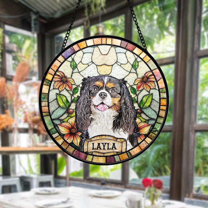 May include: A round stained glass window featuring a Cavalier King Charles Spaniel with a black, brown, and white coat. The dog is surrounded by floral and leaf designs in shades of orange, green, and yellow. The name "LAYLA" is displayed on a banner.