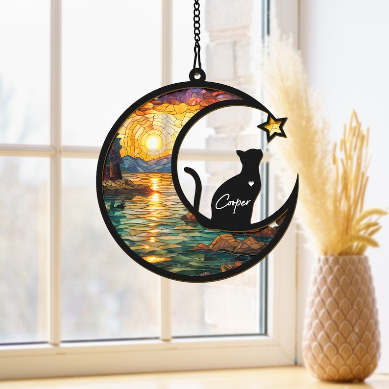 May include: A black cat silhouette with the name "Cooper" is sitting on a crescent moon with a stained glass sunset scene. The moon is hanging from a chain.