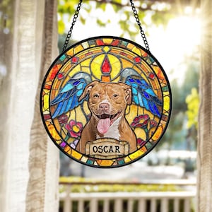 May include: Round stained glass window featuring a smiling brown dog with blue angel wings. The dog's name, "OSCAR", is displayed on a banner. The window has a black border and is suspended by a chain.