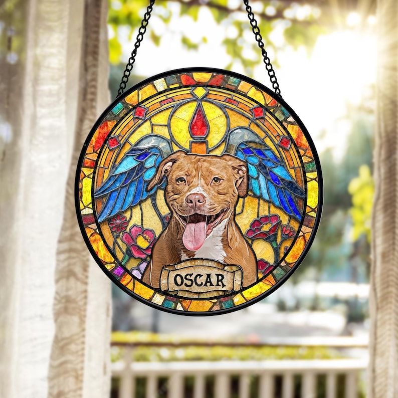 May include: Round stained glass window featuring a smiling brown dog with blue angel wings. The dog's name, "OSCAR", is displayed on a banner. The window has a black border and is suspended by a chain.