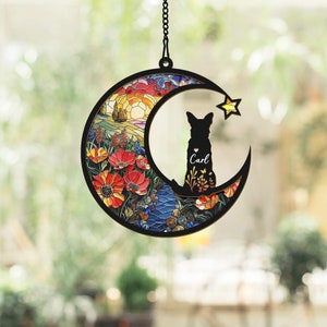 Pode incluir: Um m&oacute;bile de vento em forma de lua de vidro colorido com uma silhueta de um c&atilde;o e o nome "Carl". A lua &eacute; decorada com um design floral colorido e uma cena de nascer do sol.