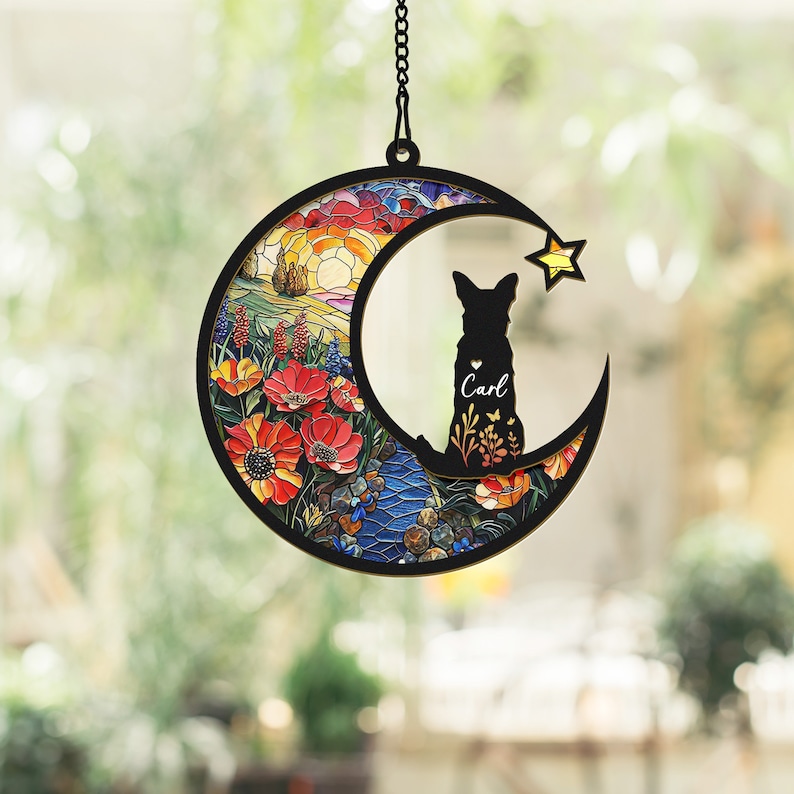 Pode incluir: Um m&oacute;bile de vento em forma de lua de vidro colorido com uma silhueta de um c&atilde;o e o nome "Carl". A lua &eacute; decorada com um design floral colorido e uma cena de nascer do sol.