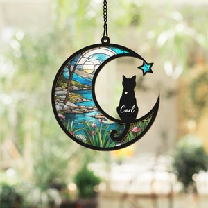 May include: A stained glass moon-shaped wind chime featuring a black cat silhouette perched on the crescent moon. The cat has a heart on its chest and the name "Carl" inscribed beneath it. The background of the wind chime depicts a colourful stained glass scene of a lake and trees.