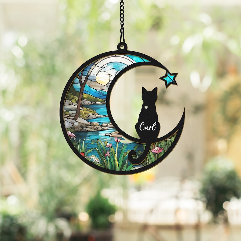 May include: A stained glass moon-shaped wind chime featuring a black cat silhouette perched on the crescent moon. The cat has a heart on its chest and the name "Carl" inscribed beneath it. The background of the wind chime depicts a colourful stained glass scene of a lake and trees.
