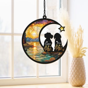 Pode incluir: Um apanhador de sol de vidro colorido com dois c&atilde;es sentados em uma lua crescente com uma cena de p&ocirc;r do sol e lago. Os c&atilde;es est&atilde;o em silhueta e t&ecirc;m os nomes "Cooper" e "Brian" escritos abaixo.
